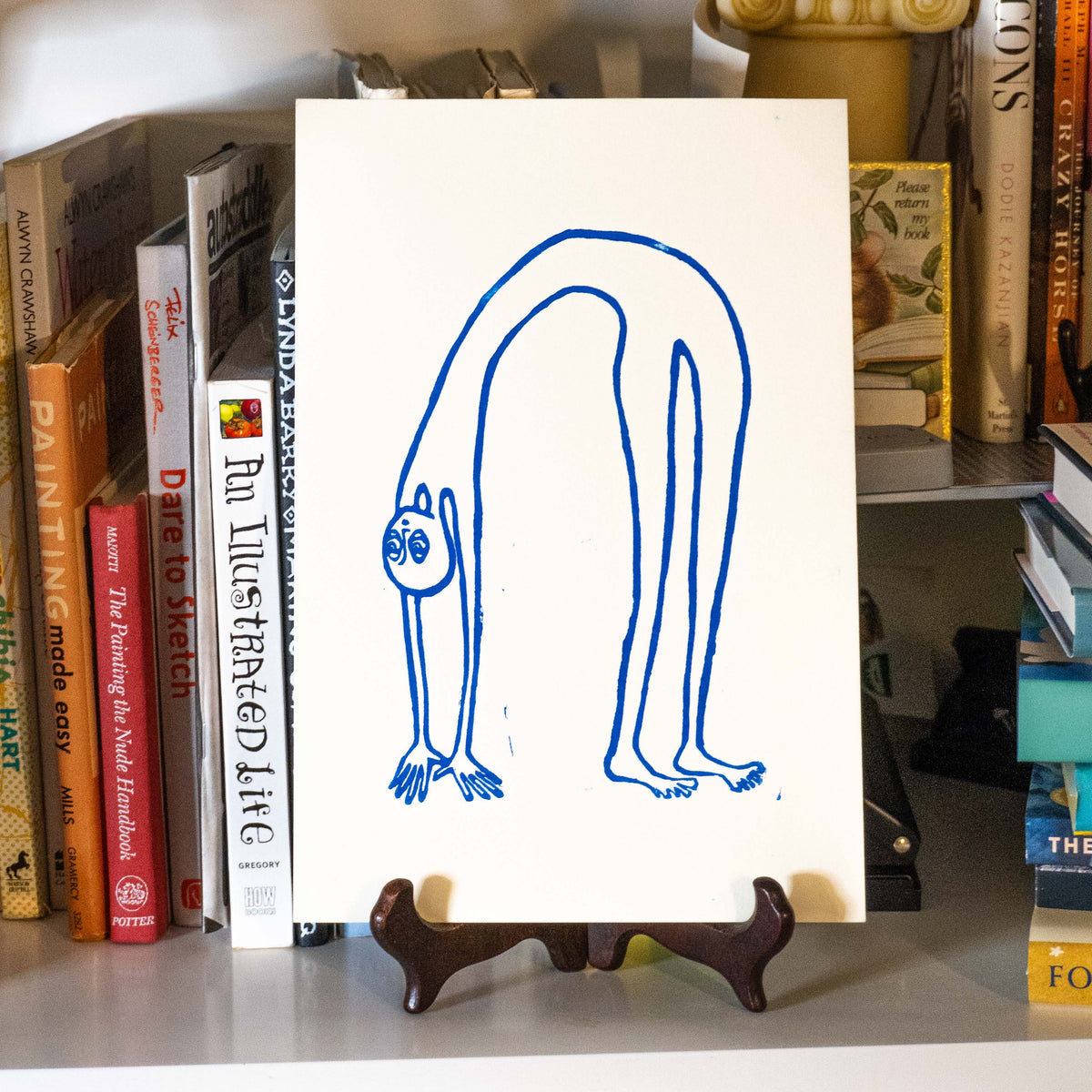 Bendy guy linocut print displayed on a wooden easel in a bookshelf.