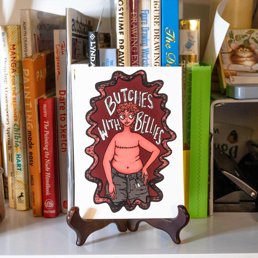 A small rectangular art print featuring an illustrated figure with top surgery scars, belly, and caribeener on jean shorts with the text "Butches with bellies" around them. Displayed on a small stand on a bookshelf.