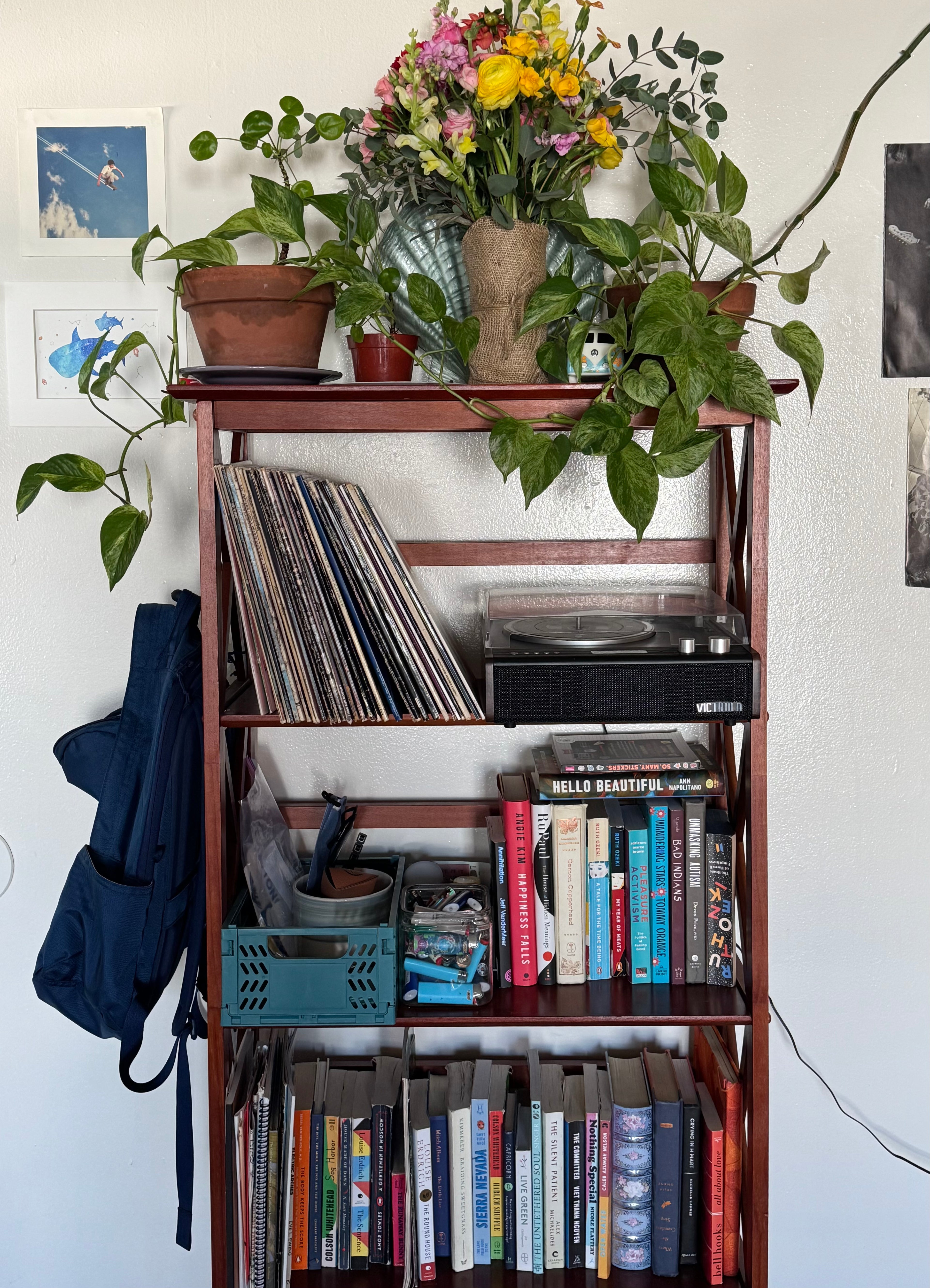 Kat’s bookshelf topped with flowers and pothos plants, holding a record player, books, records, a container of lighters, and a basket of keys and sunglasses. A blue backpack hangs on one side, and the wall behind is covered in pictures and prints.