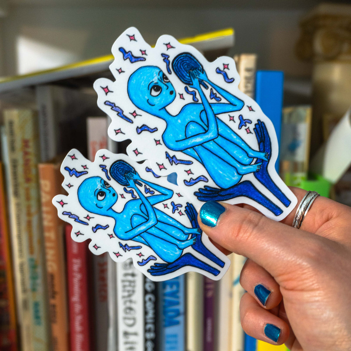 A hand with blue nail polish holds both size variations of the alien mirror sticker in front of a blurred bookshelf.