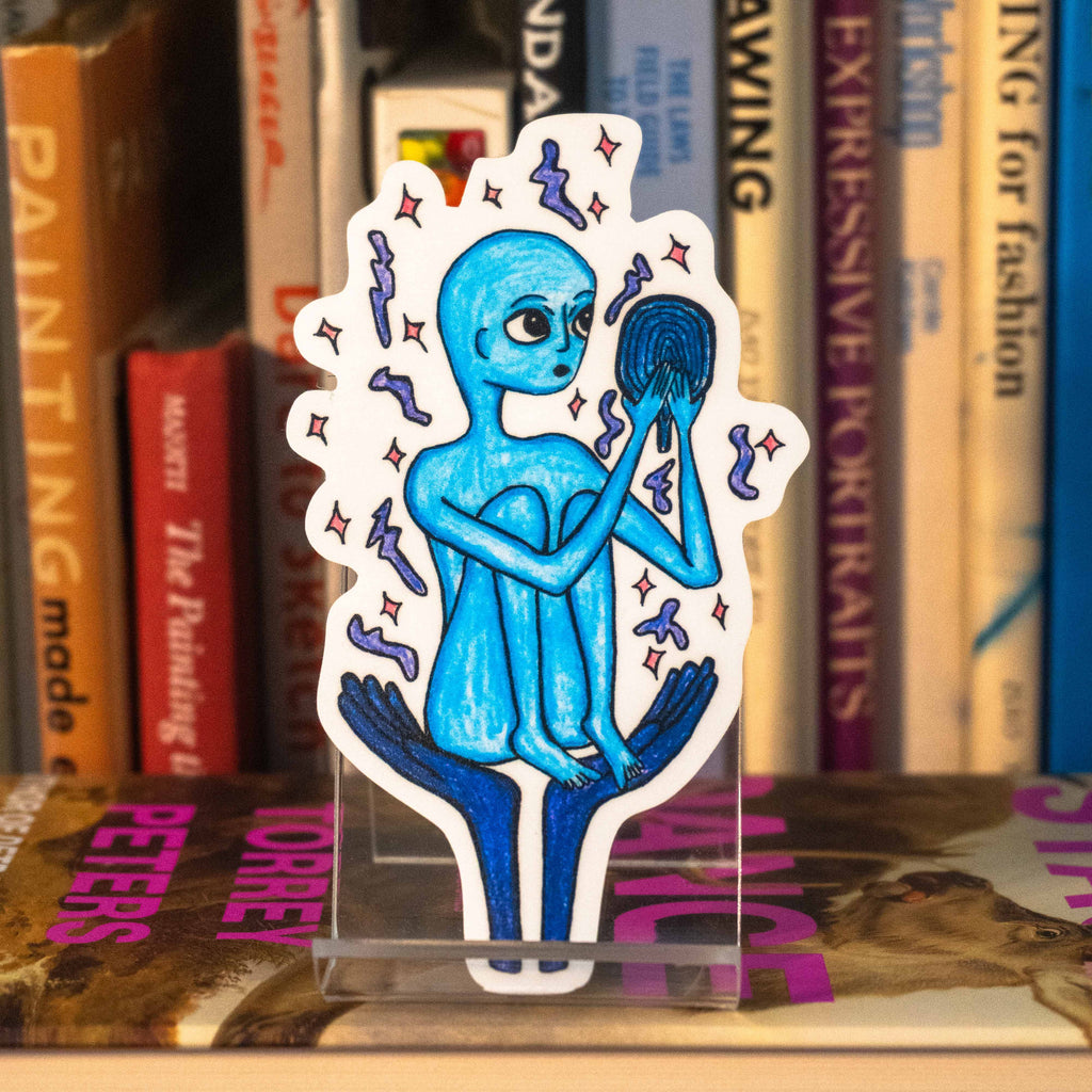 A large die-cut sticker featuring a blue alien holding a mirror, displayed on a clear acrylic stand in front of a bookshelf.