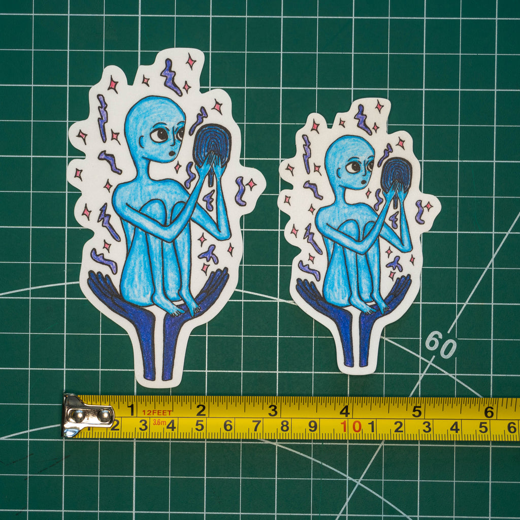 Two differently-sized die-cut stickers featuring a blue alien holding a mirror placed on a green cutting mat with a yellow tape measure below for scale.