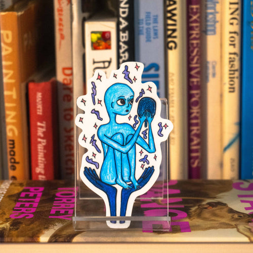 A small die-cut sticker featuring a blue alien holding a mirror, displayed on a clear acrylic stand in front of a bookshelf.