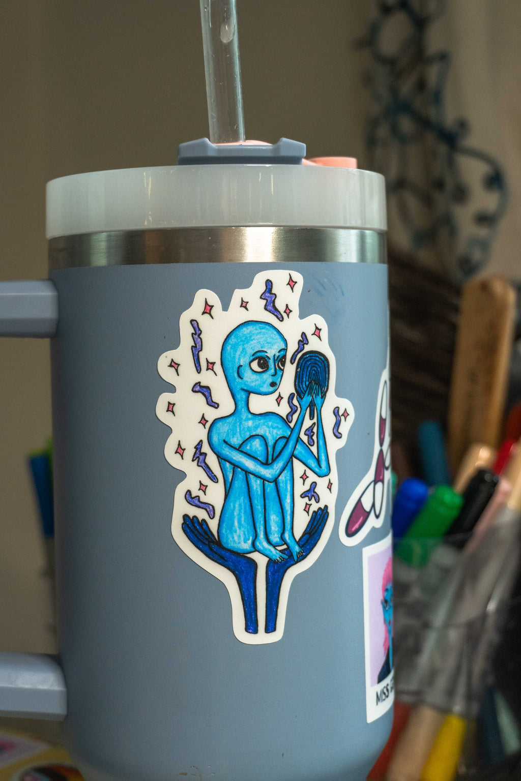 A gray insulated tumbler with a clear straw and lid, decorated with a die-cut sticker of a blue alien holding a mirror. The tumbler is shown in a real-world setting with pens and other items blurred in the background.
