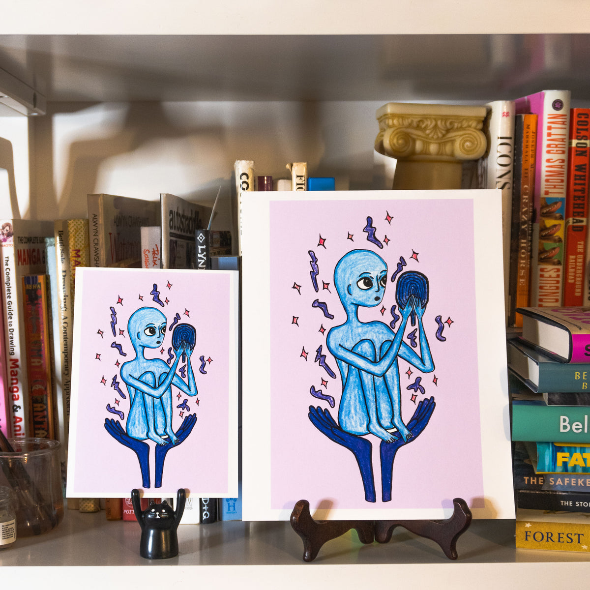 Two art prints featuring a blue alien holding a mirror, displayed on a bookshelf between books.