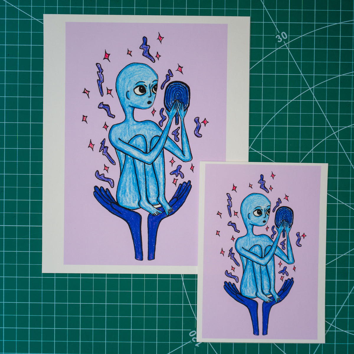 Two art prints featuring a blue alien sitting on blue hands against a lavender background, displayed on a green cutting mat. One print is larger and positioned behind the other.