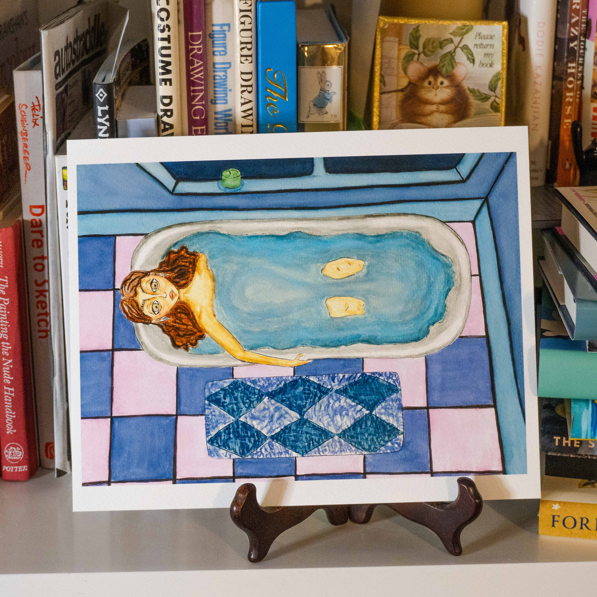 A watercolor art print depicting a woman relaxing in a blue bathtub on a pink and blue tiled floor, displayed on a small wooden easel in front of a bookshelf.