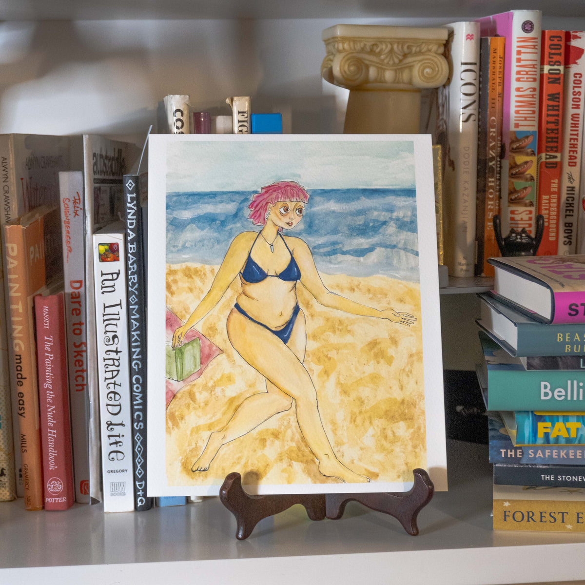 A watercolor art print of a woman with pink hair in a blue bikini walking on a sandy beach, displayed on a small wooden easel stand on a bookshelf surrounded by books.