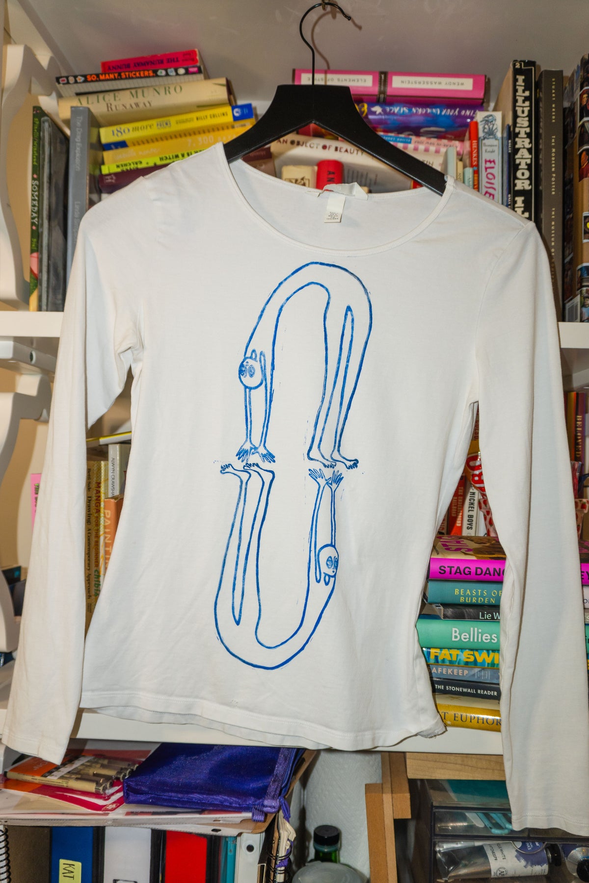 A white long-sleeve t-shirt with a blue linocut print of figures in a backbend, hanging on a black hanger in front of a bookshelf.