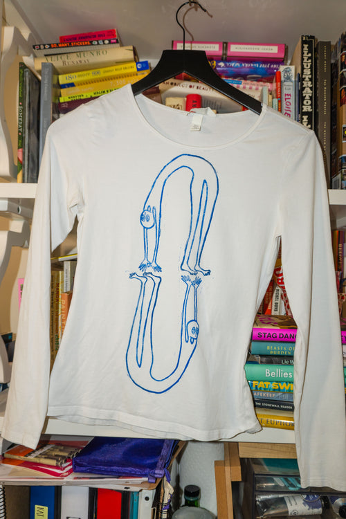 A white long-sleeve t-shirt with a blue linocut print of figures in a backbend, hanging on a black hanger in front of a bookshelf.