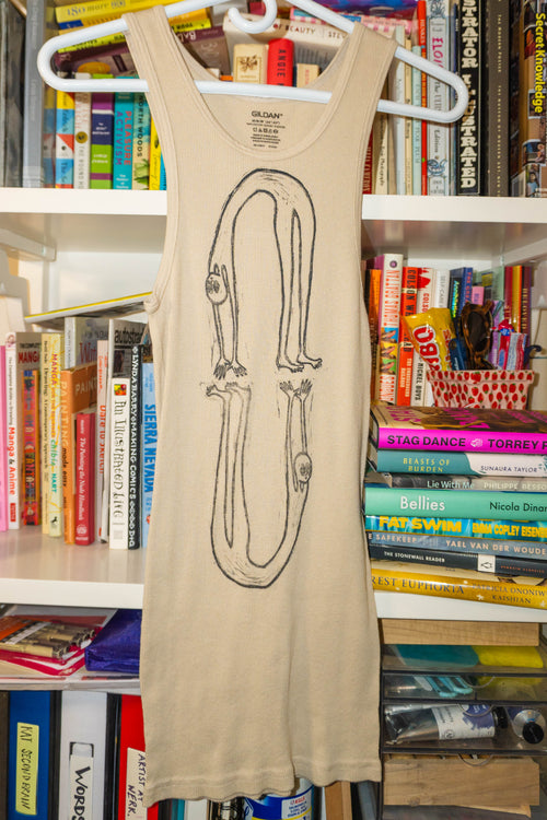 A beige sleeveless tank dress hanging on a white hanger in front of a bookshelf filled with books. The tank features a large linocut print of two figures in a backbend.