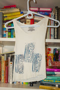 A cream-colored tank top with a blue linocut abstract graphic print hanging on a white plastic hanger, set against a background of bookshelves filled with books.