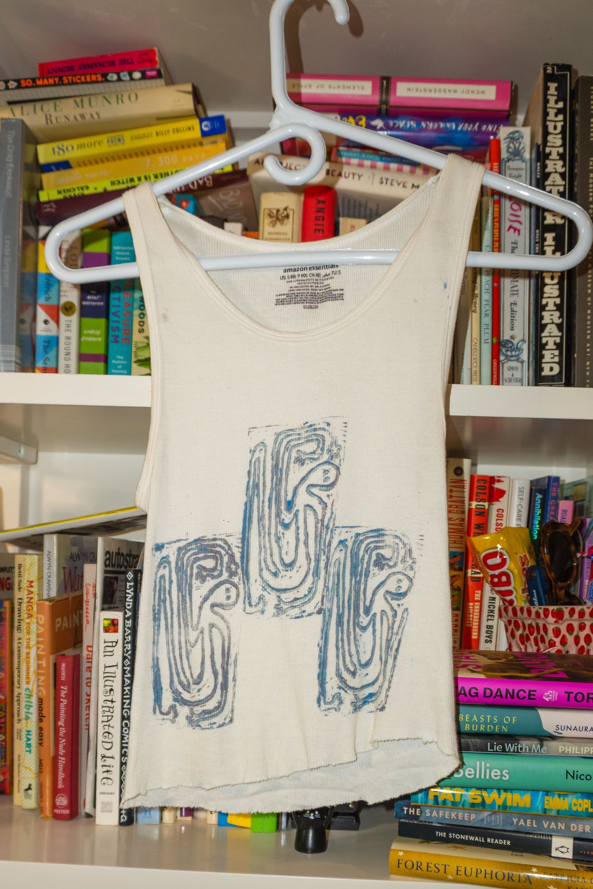 A cream-colored tank top with a blue linocut abstract graphic print hanging on a white plastic hanger, set against a background of bookshelves filled with books.