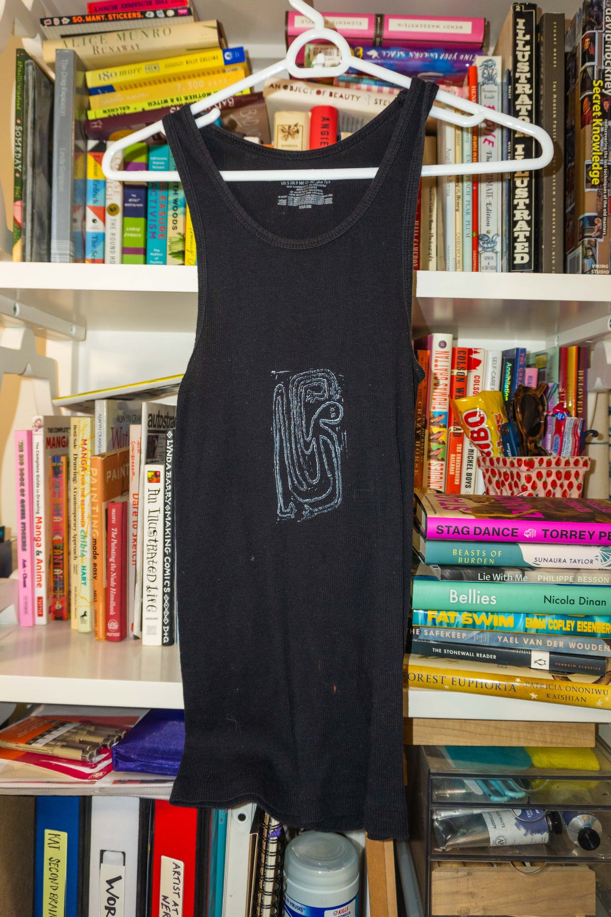 A black sleeveless tank top with a stylized gray linocut print on the front, hanging on a white plastic hanger in front of a bookshelf filled with books.