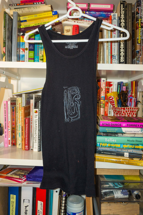 A black sleeveless tank top with a stylized gray linocut print on the front, hanging on a white plastic hanger in front of a bookshelf filled with books.