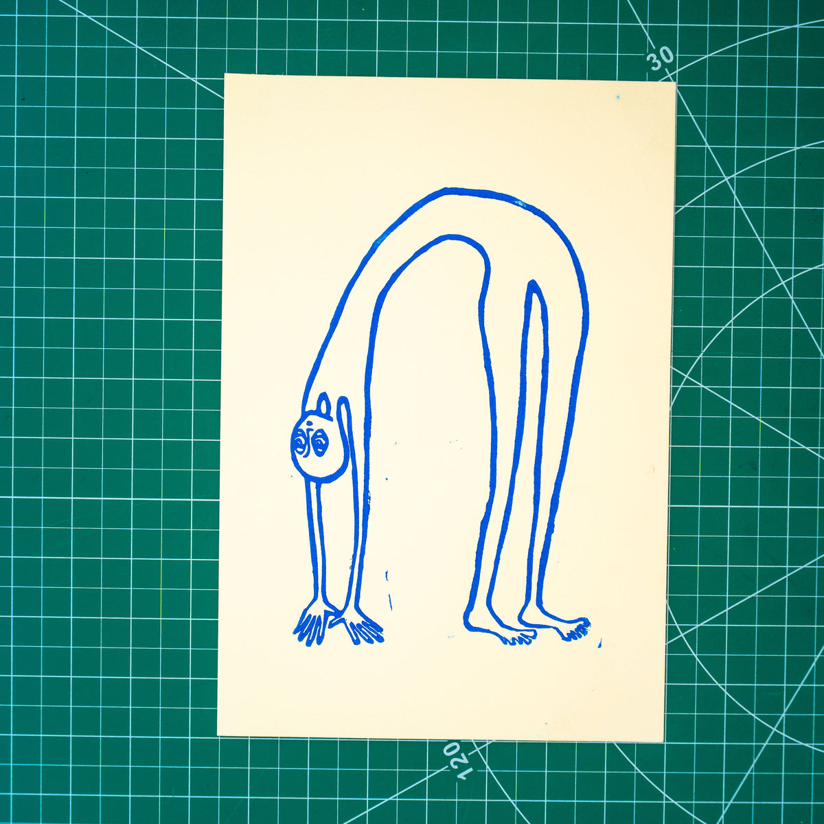 A rectangular white art print featuring a blue linocut design of a alien-like figure in a backbend, displayed on a green cutting mat with grid lines