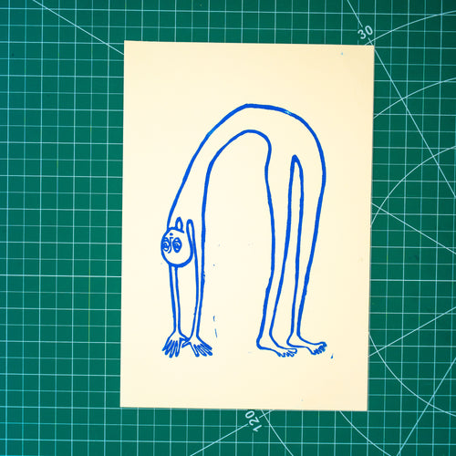 A rectangular white art print featuring a blue linocut design of a alien-like figure in a backbend, displayed on a green cutting mat with grid lines