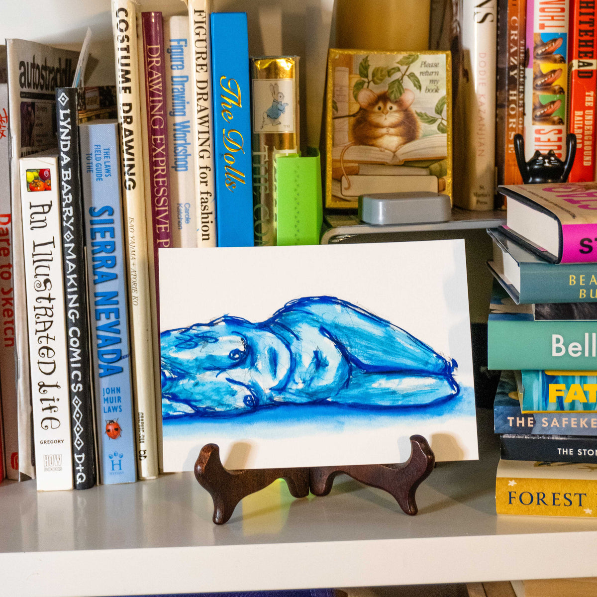 A small art print or card featuring a blue watercolor-style drawing of a sleeping figure, displayed on a small wooden easel stand on a bookshelf surrounded by books.