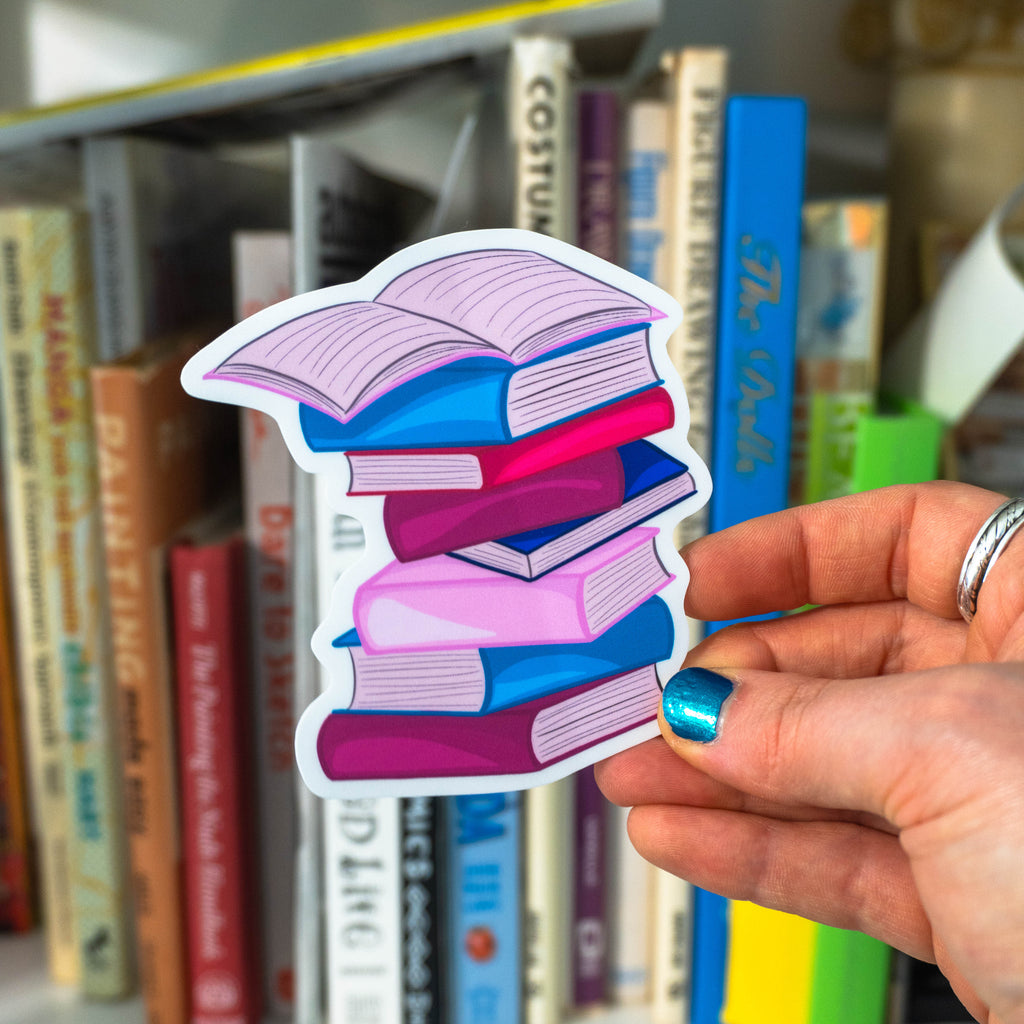 A hand with blue glitter nail polish holds a large die-cut sticker shaped like a stack of colorful open books, with a blurred background of bookshelves.