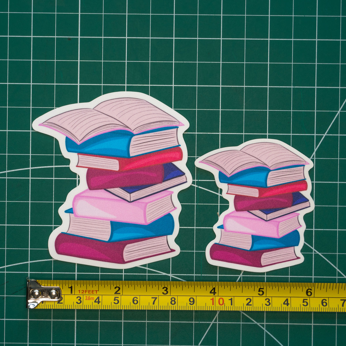 Two die-cut stickers depicting stacks of colorful books in different sizes, shown on a green cutting mat with a yellow tape measure for scale.