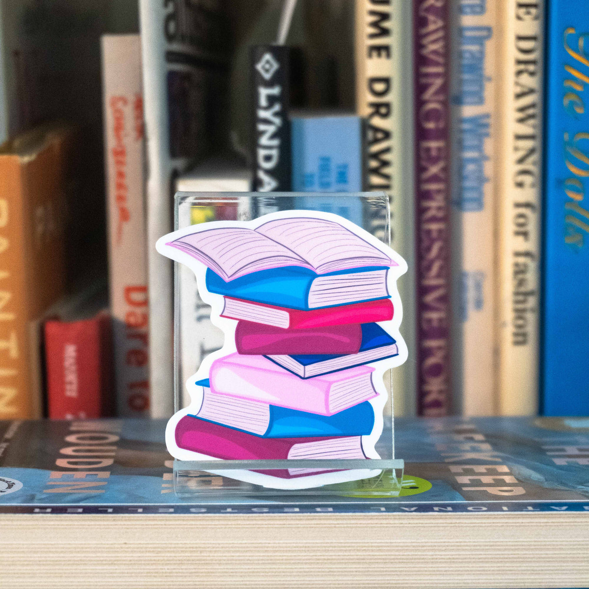 A small die-cut sticker of a stack of colorful books, displayed on a clear acrylic stand on a bookshelf in front of blurred books.