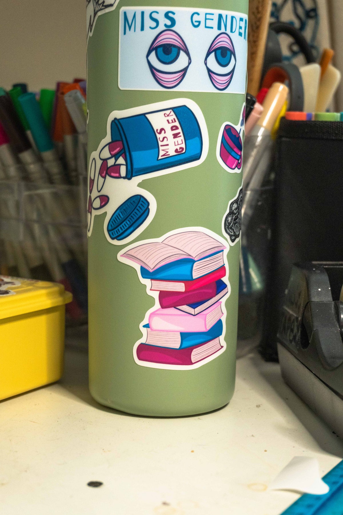 A sage green insulated tumbler with stickers, including a 'Miss Gender' eye sticker and a book stack sticker, shown on a desk with office supplies in the background.