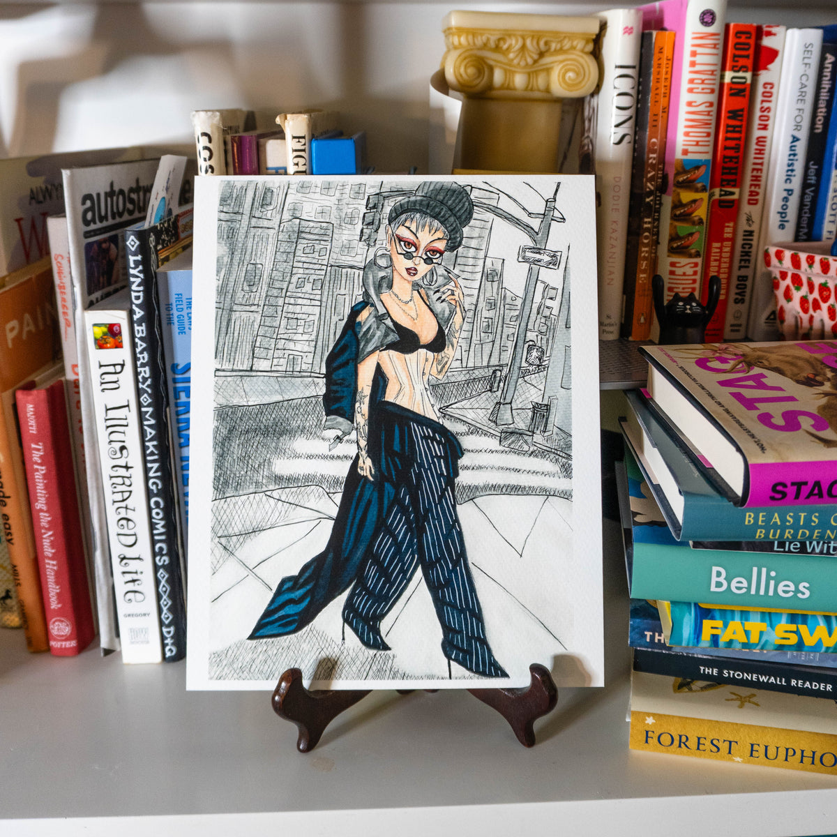 A black and white art print featuring a stylized illustration of a woman in a black outfit, displayed on a small wooden easel stand in front of a bookshelf filled with books.
