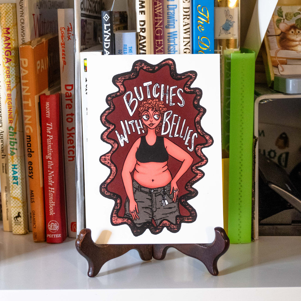 A small rectangular art print featuring an illustrated figure with a black sports bra, belly, and caribeener on jean shorts with the text "Butches with bellies" around them. Displayed on a small stand in a bookshelf.