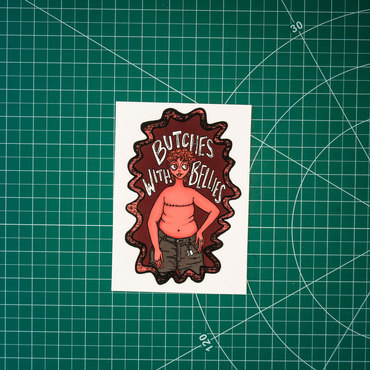 A small rectangular art print featuring an illustrated figure with top surgery scars, belly, and caribeener on jean shorts with the text "Butches with bellies" around them, displayed on a green cutting mat with grid lines.