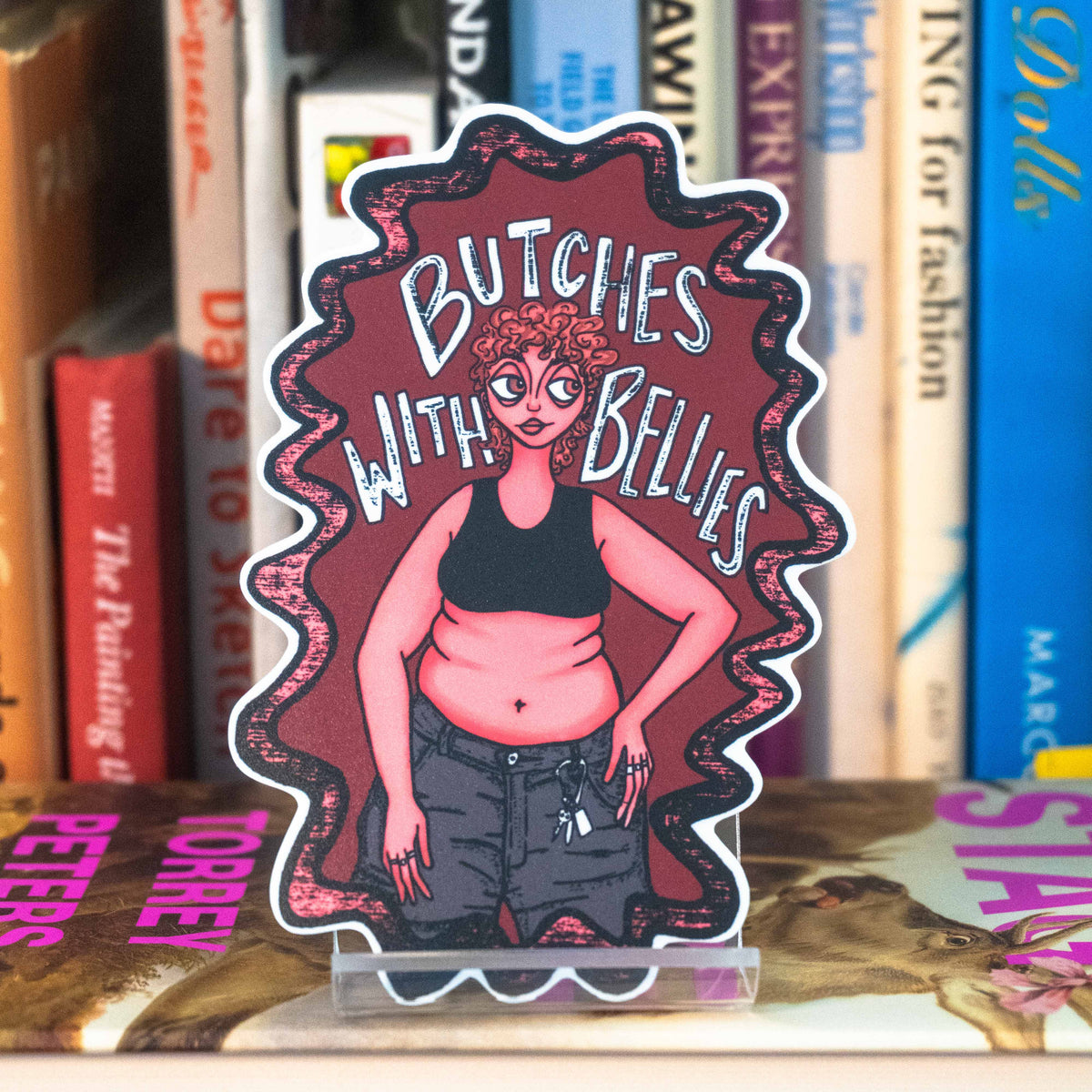 A large die-cut sticker featuring an illustrated figure with a black sports bra, belly, and caribeener on jean shorts with the text "Butches with bellies" around them. displayed on a clear acrylic stand in front of a bookshelf.