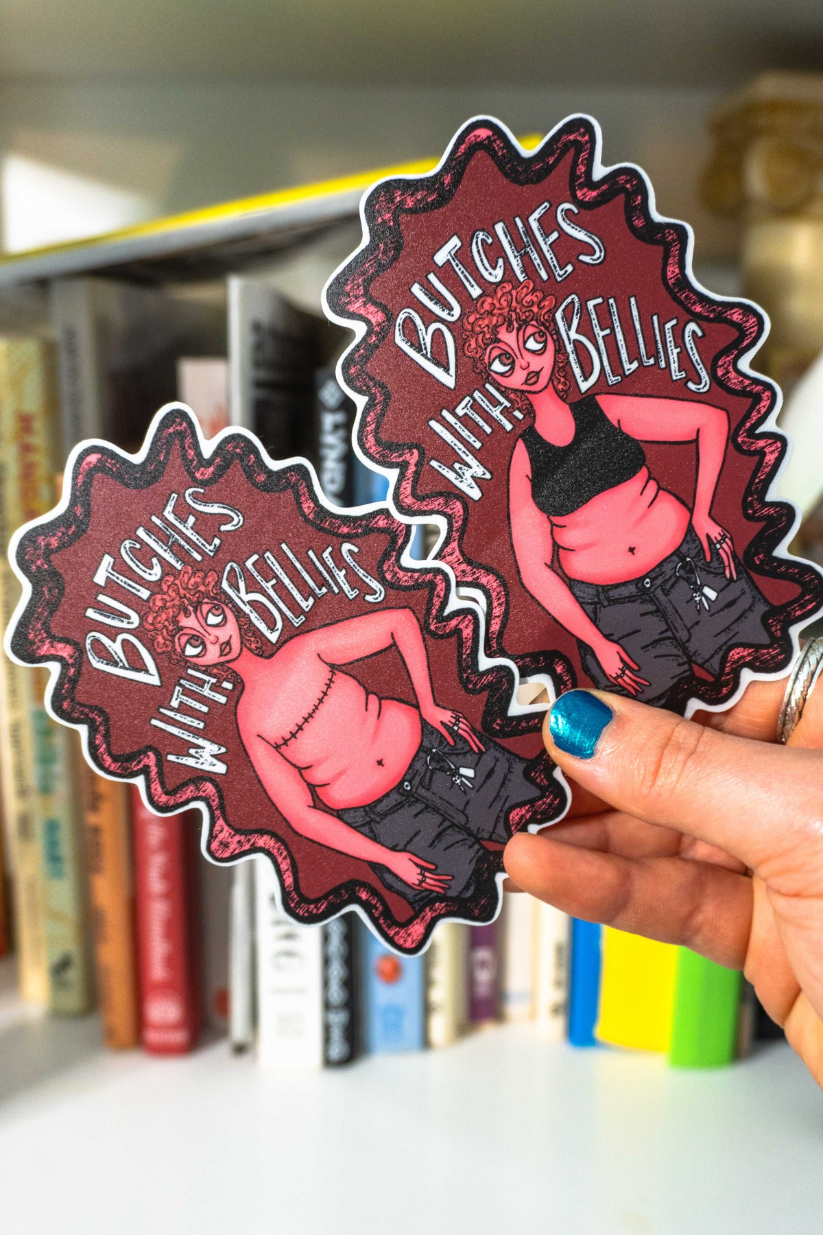 Hand with blue nail polish holding both variations of the "Butches with bellies" sticker in front of a blurred bookshelf.
