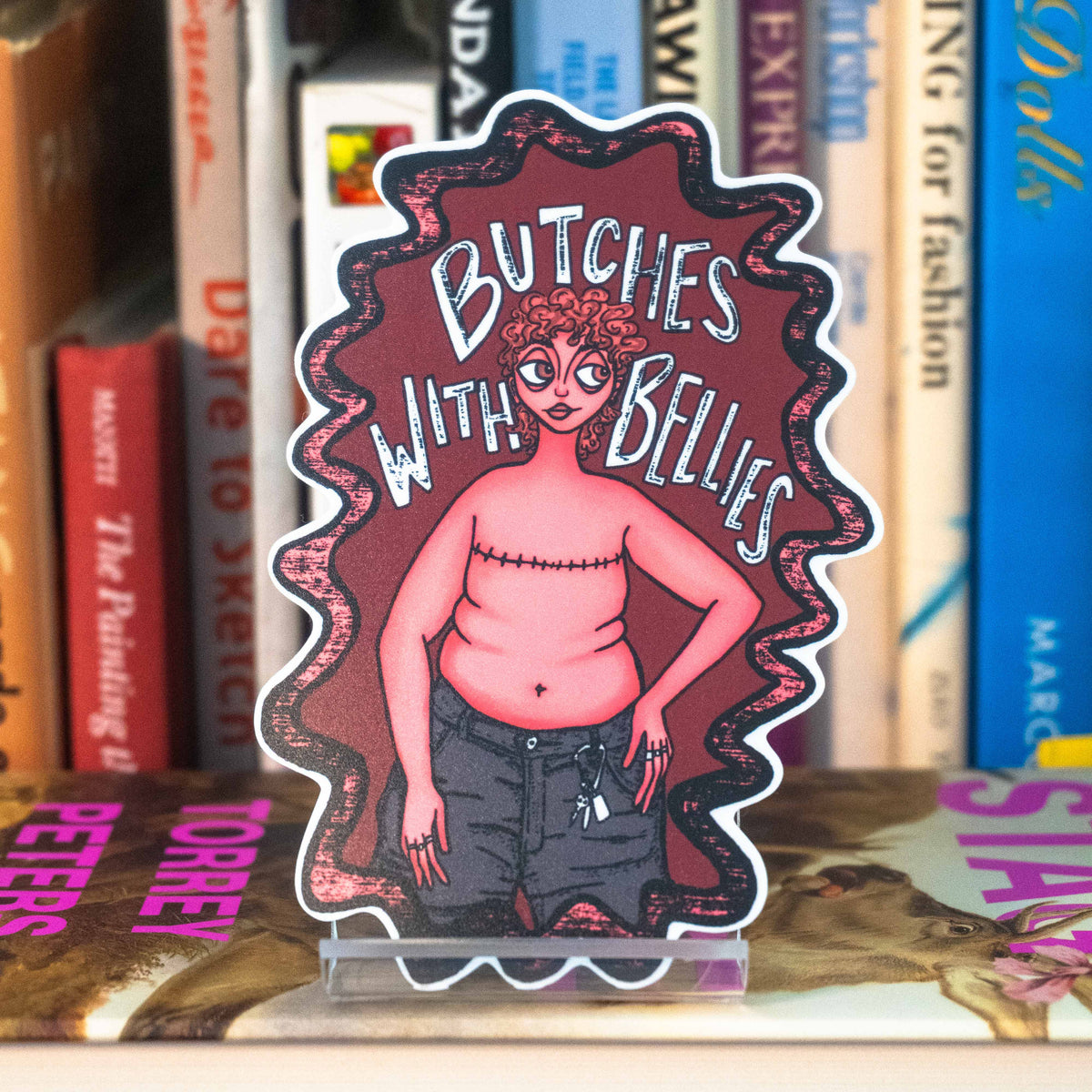 A large die-cut sticker featuring an illustrated figure with top surgery scars, belly, and caribeener on jean shorts with the text "Butches with bellies" around them. displayed on a clear acrylic stand in front of a bookshelf.