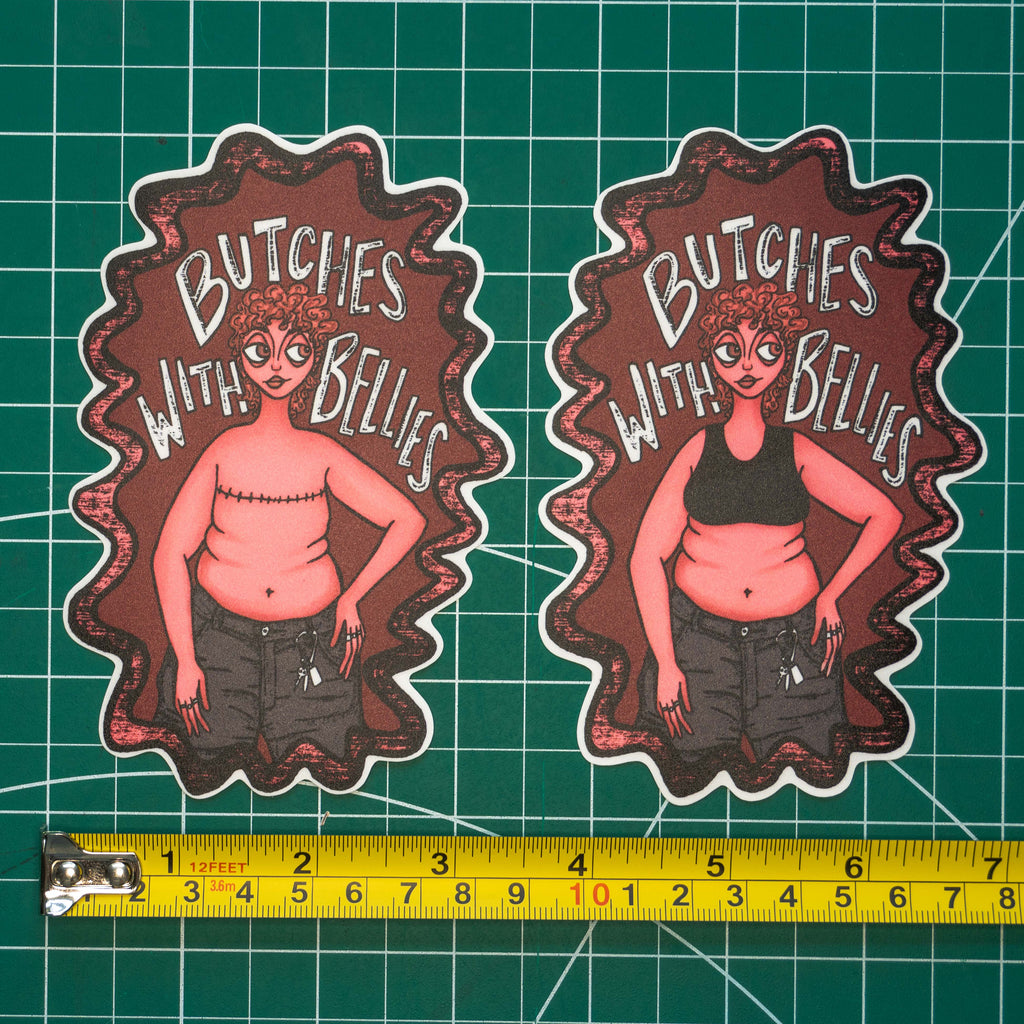 Two large die-cut stickers featuring an illustrated figure with the text "Butches with bellies" around them, displayed on a green cutting mat with a tape measurer for scale. The sticker on the right depicts the figure in a sports bra while the one on the left shows the figure with top surgery scars across their chest. 