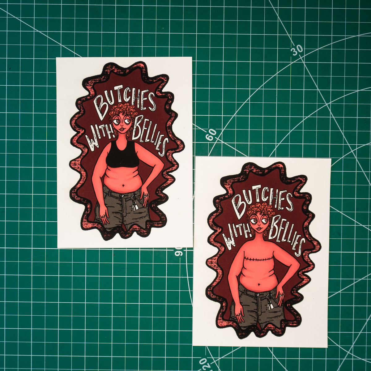 Two small rectangular art prints featuring an illustrated figure with the text "Butches with bellies" around them. The art print on the left depicts the figure in a sports bra while the one on the right shows the figure with top surgery scars across their chest. Displayed on a green cutting mat.