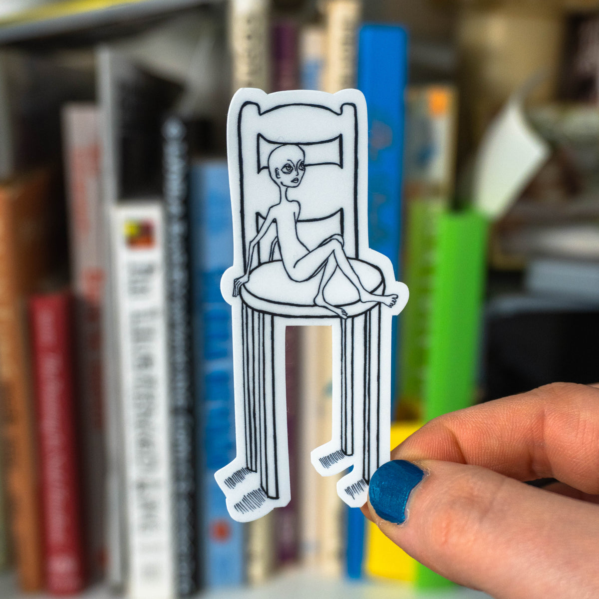 Hand with blue nail polish holding die-cut little guy in chair sticker in front of a blurred bookshelf.