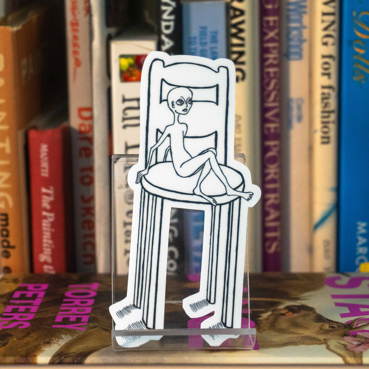 A large die-cut sticker featuring an illustration of an alien-like figure sitting in a disproportionately large chair, displayed on a clear acrylic stand in front of a bookshelf.