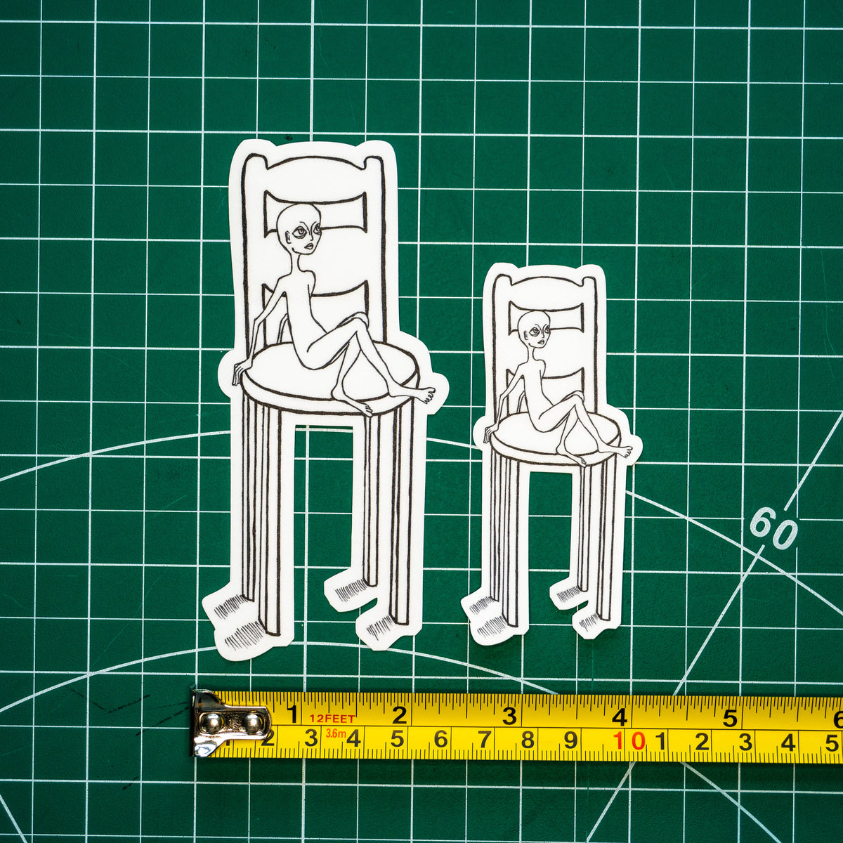 Two differently-sized die-cut stickers featuring an illustration of an alien-like figure sitting in a disproportionately large chair placed on a green cutting mat with a yellow tape measure below for scale.