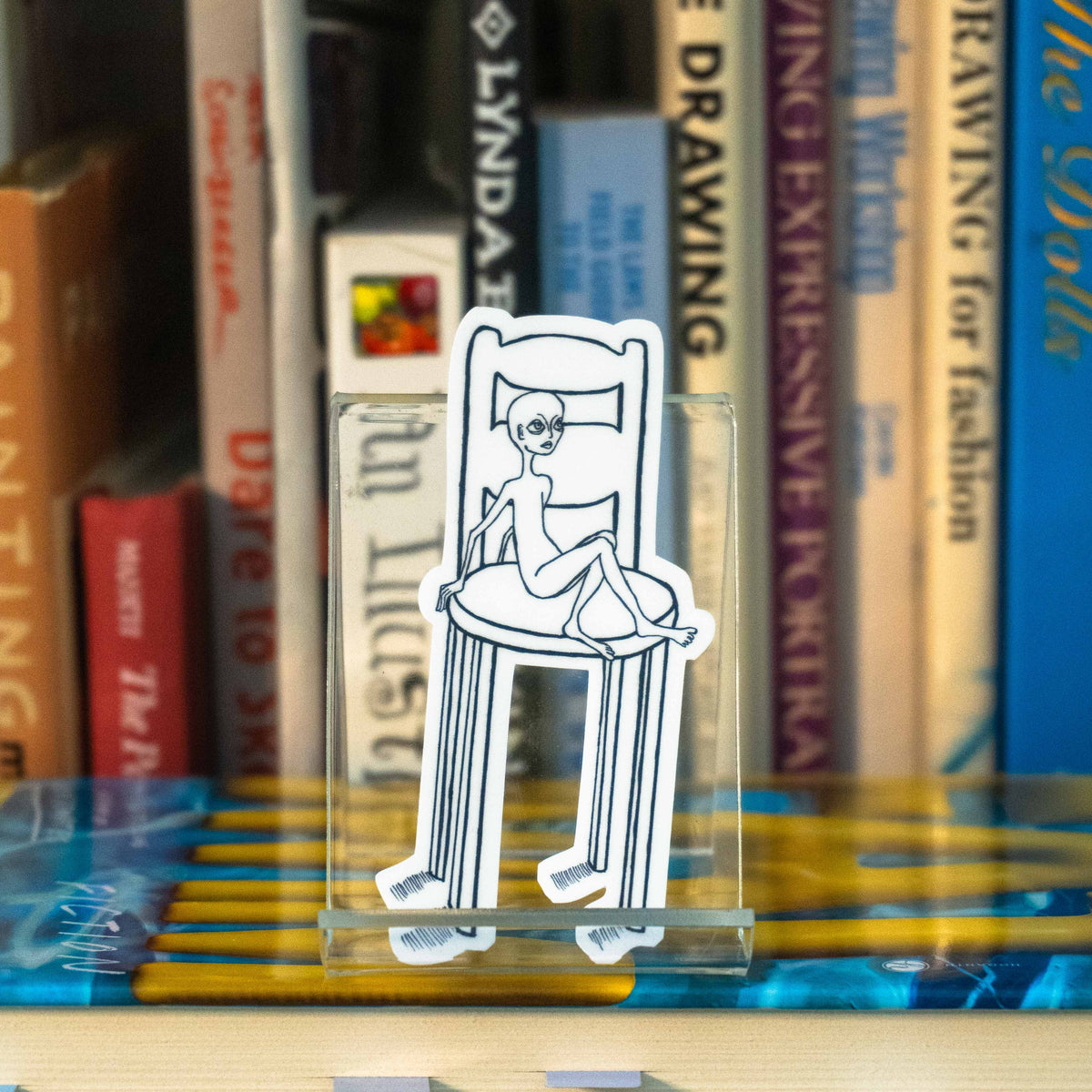 A small die-cut sticker featuring an illustration of an alien-like figure sitting in a disproportionately large chair, displayed on a clear acrylic stand in front of a bookshelf.