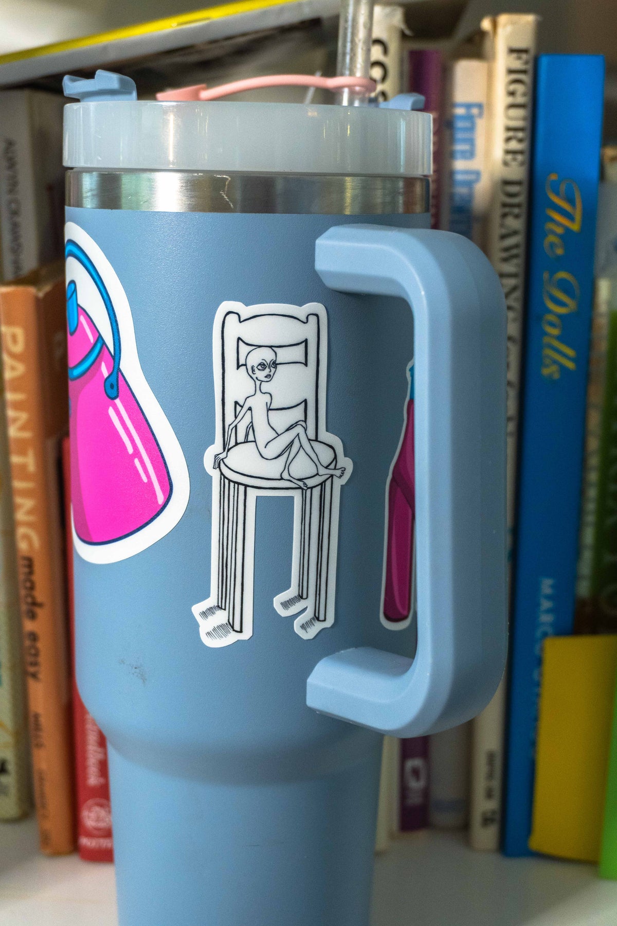 A light blue insulated travel tumbler with a handle, a clear lid, and a pink straw, decorated with die-cut sticker of little guy in a big chair. 