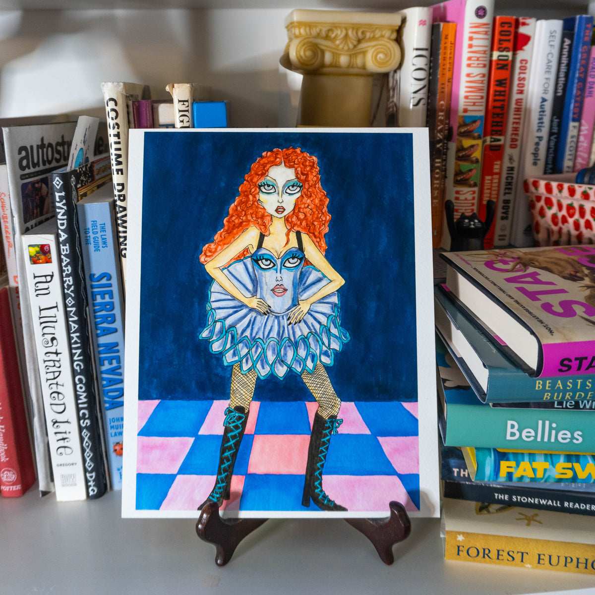 A square art print featuring a stylized illustration of a woman with bright red hair, a blue tutu, and patterned boots, displayed on a small stand in front of a bookshelf.