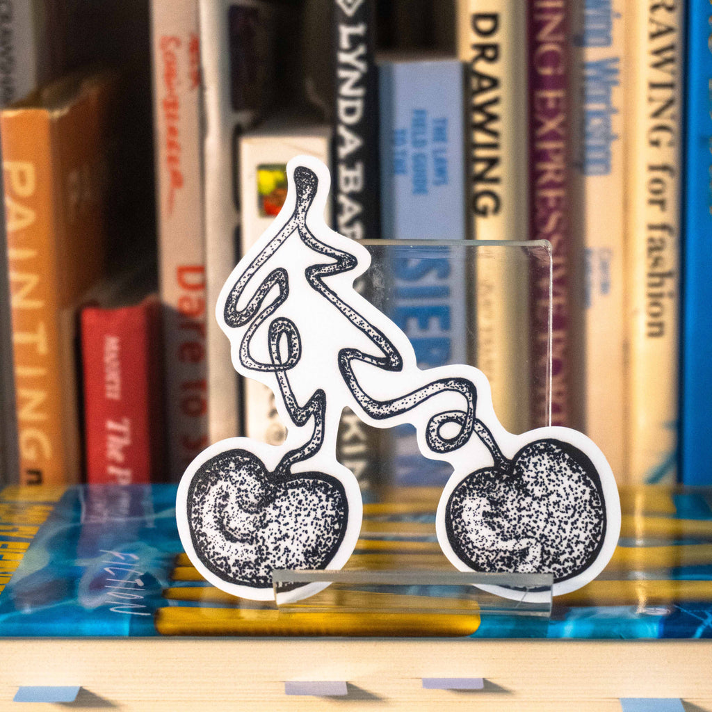 A large die-cut sticker featuring a grayscale tattoo-style illustration of a cherry displayed on a clear acrylic stand in front of a bookshelf.
