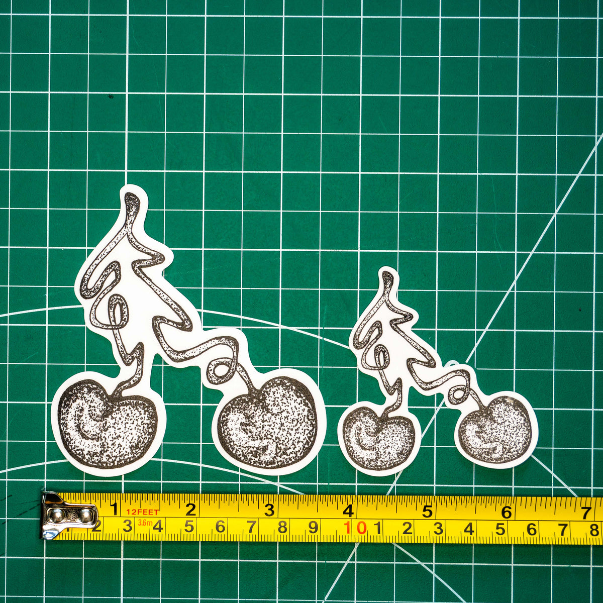 Two differently-sized die-cut stickers featuring a grayscale tattoo-style illustration of a cherry displayed on a green cutting mat with a tape measure for scale.
