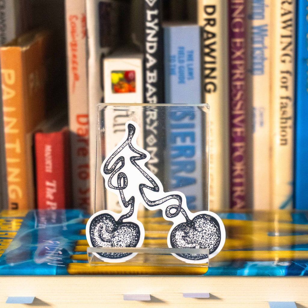 A small die-cut sticker featuring a grayscale tattoo-style illustration of a cherry displayed on a clear acrylic stand in front of a bookshelf.