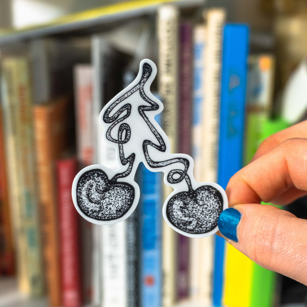 A hand with blue nail polish holds small cherry die-cut sticker in front of a background of blurred books. 