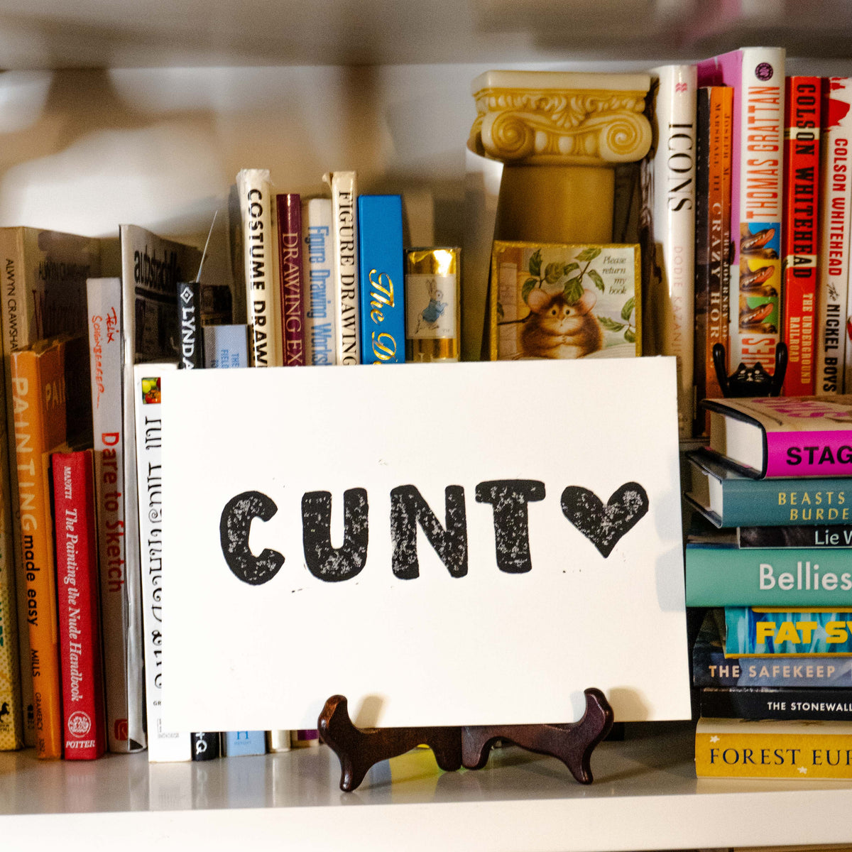 "Cunt <3" linocut print displayed on easel in bookshelf.