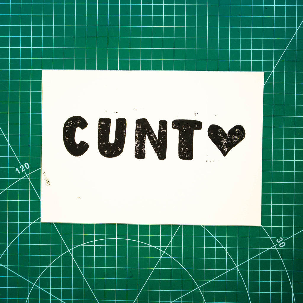 A white rectangular print featuring "cunt <3" lino block design, displayed on green cutting mat with grid lines.
