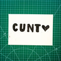 A white rectangular print featuring "cunt <3" lino block design, displayed on green cutting mat with grid lines.