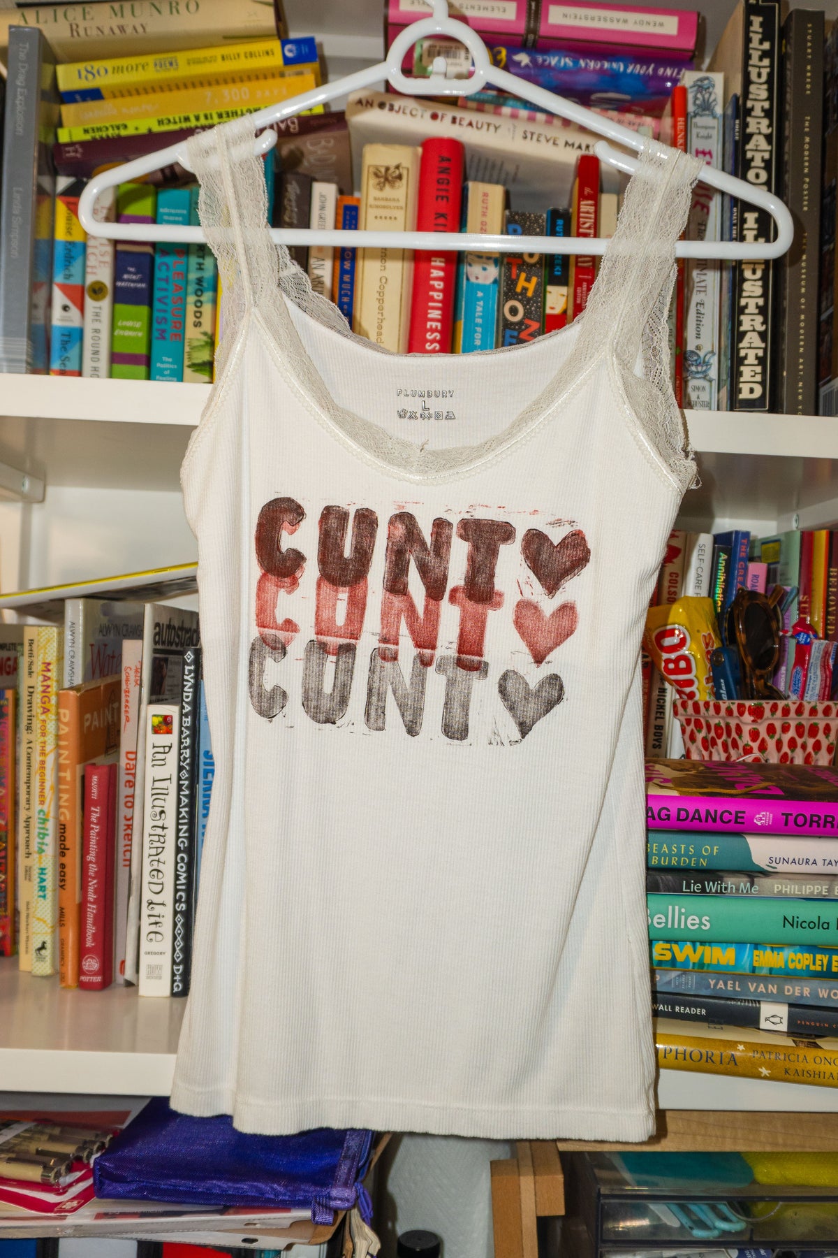 A white sleeveless tank top with "cunt <3" linocut print in shades of red and black across the chest, hanging on a white hanger in front of a bookshelf.