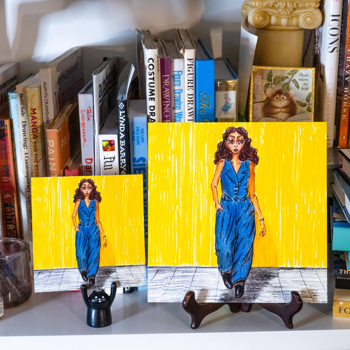 Two hand-drawn art prints of different sizes depicting a woman in a blue denim suit against a bright yellow background, displayed on a bookshelf.