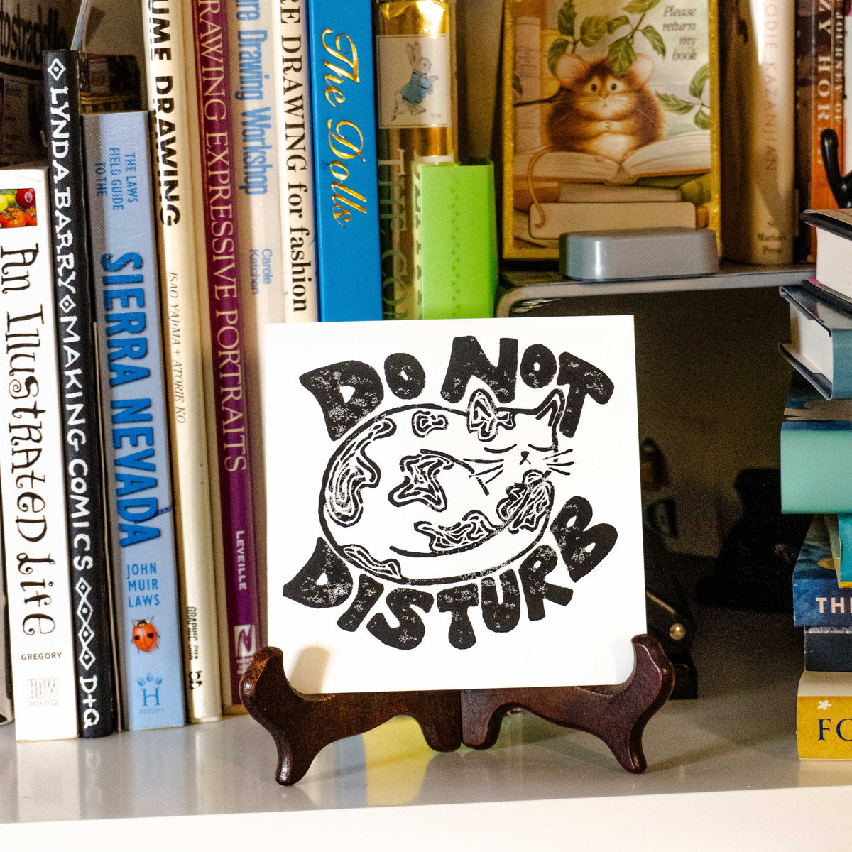 "Do Not Disturb" lino print displayed within shelf filled with tsachkis and books.